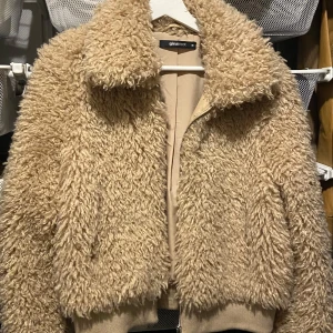 Beige teddyjacka från Gina Tricot - Mysig beige teddyjacka från Gina Tricot med dragkedja. Perfekt för kyliga dagar med sin fluffiga och varma design. Jackan har en klassisk krage och ribbade muddar vid ärmsluten.