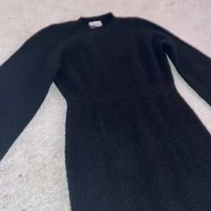 Svart stickad klänning från Aware - Elegant svart stickad klänning från Aware med lång ärm och en stilren design. Klänningen har en smickrande passform och en subtil slits i nederkanten. Perfekt för kyligare dagar.