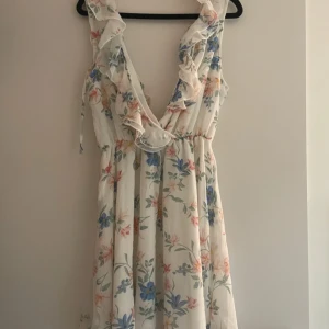 Blommig klänning från H&M - Charmig blommig klänning från H&M i vitt med färgglada blommor. Klänningen har volangdetaljer och en v-ringad design. Perfekt för sommarens varma dagar.