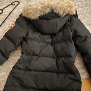 Svart dunjacka med päls från Canada Goose - Säljer en svart dunjacka från Canada Goose med en mysig pälsfodrad huva. Jackan har en klassisk design med lång ärm och är perfekt för kalla vinterdagar. Den har en dragkedja framtill och en logotyp på ärmen. Är inte i behov av pengarna vill bara bli av med den.