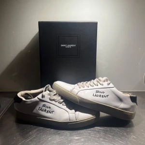 Vita sneakers från Saint Laurent - Snygga vita sneakers från Saint Laurent med handskriven logga på sidan. Skorna har en klassisk design med snörning och en bekväm sula. Perfekta för en stilren look. Skorna har en del defekter som man kan se på bilderna. Fråga om du vill ha tydligare bilder. Säljer för dunderpris pga detta och är osäker på äktheten för jag köpte de på plick. //Tom🦁