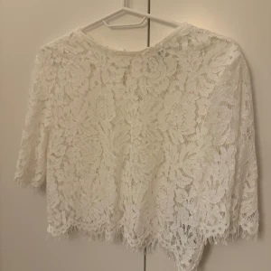 Vit spetstopp - Säljer en elegant vit spetsblus med trekvartsärm och vackert blommönster. Blusen har en rund hals och en liten knäppning i nacken. Perfekt för en stilren look.