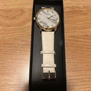Klocka med marmormönster - Klocka med vit/beige läderrem och urtavla i marmormönster. Klockan har romerska siffror och guldfärgade detaljer. Helt ny, ej använd och fortfarande i orginal kartong. Byte av batteri kan behövas då det troligtvis laddat ur💗