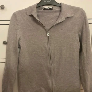 Grå kofta från J.Lindeberg i kashmire  - Säljer en stilren grå kofta från J.Lindeberg med dragkedja framtill. Perfekt för lager-på-lager under kyligare dagar. Koftan har en klassisk krage och långa ärmar.