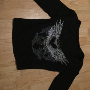 Svart tröja med vingmönster - Snygg svart tröja med ett coolt tryck av ett hjärta med vingar i grått och vitt. Tröjan har långa ärmar och en bekväm passform.