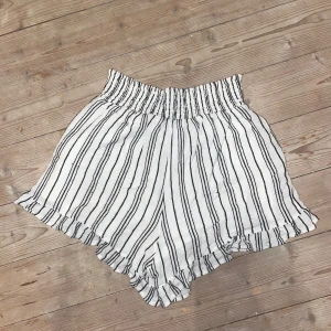 Randiga shorts från KappAhl - Snygga vita shorts med svarta ränder från KappAhl. De har en elastisk midja och volangdetaljer vid bensluten, vilket ger en avslappnad och trendig look. Perfekta för varma sommardagar!