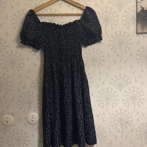 Mörkblå klänning med prickar - Charmig mörkblå klänning med vita prickar och puffiga kortärmar. Den har en smockad överdel som ger en bekväm passform och en luftig kjol. Perfekt för en somrig look!