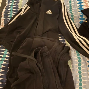 Svart träningsjacka och byxor från Adidas - Snygg svart träningsjacka från Adidas med de klassiska tre vita ränderna längs ärmarna. Jackan har dragkedja och Adidas-logga på bröstet. Perfekt för sport och fritid. Matcha med de svarta byxorna med hel svarta ränder för en perfekt match