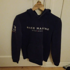 Mörkblå hoodie från Race Marine - Säljer en snygg mörkblå hoodie från Race Marine med tryck på bröstet och ärmen. Perfekt för en avslappnad stil med skandinavisk touch. Hoodien har en klassisk passform och är tillverkad i mjukt material.