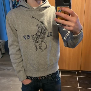 Ralph Lauren hoodie - Säljer en grå hoodie från Ralph Lauren med ett stort broderat raffe märke. Tröjan har en klassisk huva och ribbade muddar vid ärmslut och nederkant. Perfekt för en avslappnad stil. Storlek XL i barn storlek så ungefär xs i vanlig storlek