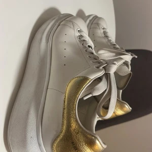 Vita sneakers från Alexander McQueen - Snygga vita sneakers från Alexander McQueen med gulddetaljer på hälen. Skorna har klassisk snörning och en tjock sula för extra komfort. Perfekta för en stilren look. Snörena är lite smutsiga men går att rengöra! Nypris 6000kr (pris kan diskuteras) 