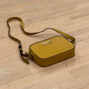 Gul axelväska från Guess - Snygg gul axelväska från Guess med präglat mönster och justerbar axelrem. Väskan har en dragkedja upptill och Guess-loggan i metall på framsidan. Perfekt för att ge en färgklick till din outfit! Endast: 299