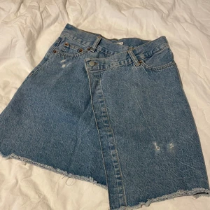 Jeanskjol från Junkyard - Snygg jeanskjol i blå denim med rå kant och slitna detaljer. Kjolen har en asymmetrisk design framtill och klassiska femficksmodellen. Perfekt för en avslappnad stil.