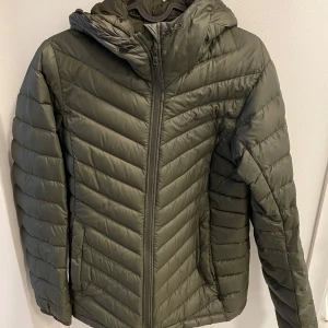 Grön pufferjacka från Peak Performance - Snygg grön pufferjacka från Peak Performance med huva och dragkedja. Perfekt för kyliga dagar med sin quiltade design som håller värmen. Jackan har en stilren look och passar bra till vardags.