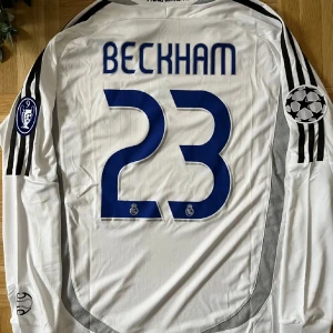 Vit Real Madrid fotbollströja med Beckham 23 - Vit långärmad Real Madrid tröja från Adidas med Beckham och nummer 23 på ryggen. Tröjan har svarta detaljer och Champions League-märke på ärmen. Framsidan pryds av BenQ Siemens-logga och klubbmärket.