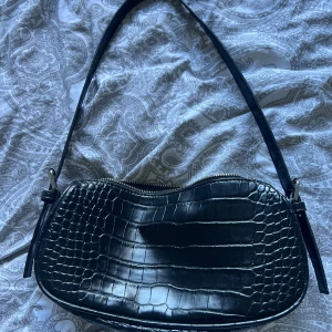 Svart axelväska i skinn - Säljer en snygg svart axelväska i skinn med krokodilpräglat mönster. Väskan har en dragkedja upptill och en justerbar axelrem. Perfekt för både vardag och fest! 🖤