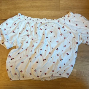 Vit off-shoulder topp med blommönster från H&M - Säljer en superfin vit off-shoulder topp från H&M. Den har ett gulligt blommönster i rosa och svart. Toppen är elastisk vid axlarna och midjan. 