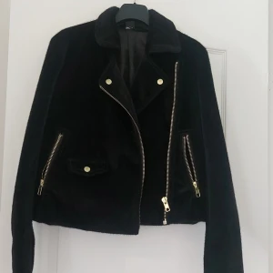 Svart mockajacka med dragkedjor - Säljer en snygg svart mockajacka med coola, guldiga dragkedjedetaljer. Jackan har en asymmetrisk dragkedja framtill och två dragkedjefickor på sidorna. Den har också en liten ficka med knapp på ena sidan. Perfekt för höst och vår! 🖤