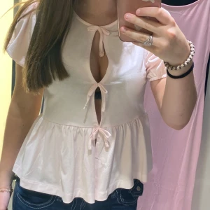 Ljusrosa puffärmad topp från Gina Tricot - Säljer en superfin ljusrosa topp från Gina Tricot. Den har puffärmar och knytband framtill som ger en söt och feminin look. Toppen är lite croppad och har en lös passform, perfekt för sommaren! Materialet känns mjukt och skönt mot huden. Använd en gång, små hål från en synål vid mitten öppningen eftersom jag sydde ihop den men har tagit bort det och märks inte alls mycket!