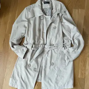 Beige trenchcoat i kortare modell, går strax ovanför knäna på mig som är 173 cm lång. Passar storlek S-M