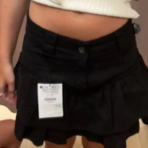 Svart kjol från Bershka - Säljer en superfin svart kjol från Bershka i storlek XS. Kjolen har en volangdetalj som gör den extra söt och perfekt för både vardag och fest. Den är gjord av ett mjukt och bekvämt material som känns skönt mot huden. Kjolen är i nyskick och har aldrig använts, prislappen sitter fortfarande kvar. Perfekt för sommaren eller en utekväll med vänner!