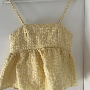 Gul blommig topp med smala axelband - Säljer en söt gul topp med smala axelband och blommigt mönster. Toppen har en smockad rygg och en lös passform nedtill. Perfekt för sommardagar!