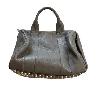 Grå handväska med nitar - Säljer min Alexander Wang Rockie bag i grått skinn med guldfärgade nitar på undersidan. Väskan har två handtag och en dragkedja för att stänga den. Perfekt för både vardag och fest! ✨Använd men i fint skick!