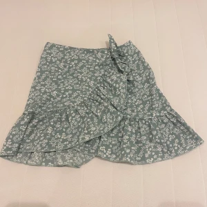 Blommig kjol med volang - Säljer en superfin blommig kjol med volangdetaljer. Kjolen är kort och har en omlott-design som ger en snygg och trendig look. Perfekt för sommaren eller en fest! 💐