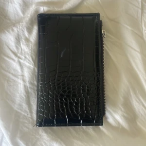 Svart plånbok med krokodilpräglat mönster - Säljer en snygg svart plånbok med krokodilpräglat mönster. Plånboken har en dragkedja på utsidan och flera fack för kort och ID på insidan. Perfekt för att hålla ordning på dina kort och kontanter. Den är i bra skick och ser väldigt stilren ut.