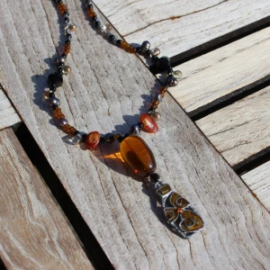 Unikt halsband med pärlor och hänge - Säljer ett unikt halsband med en mix av pärlor i olika färger och material. Halsbandet har ett stort hänge med en brun sten och ett detaljerat metallhänge med mönster. Perfekt för att ge en bohemisk touch till din outfit!