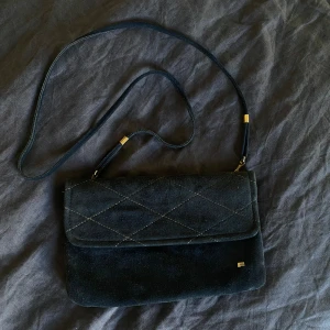 Svart quiltad axelväska - Säljer en snygg marinblå axelväska med quiltad design. Väskan är i mocka och har en praktisk dragkedja för säker förvaring. Den har en axelrem med gulddetaljer som ger en lyxig touch. Perfekt för både vardag och fest!