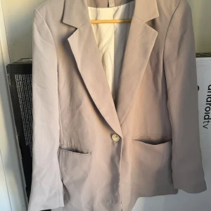 Beige kavaj från H&M - Säljer en stilren beige kavaj från H&M i storlek S. Kavajen har en klassisk design med en knapp framtill och två fickor. Perfekt för både vardag och festliga tillfällen!