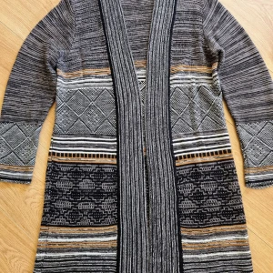 Mönstrad lång kofta från Freequent - Säljer en superfin lång kofta från Freequent i storlek . Den är stickad och har ett coolt mönster i svart, grått och beige. Perfekt för höst och vinter med sina långa ärmar och mysiga känsla. Koftan har en öppen design utan knappar, vilket gör den lä