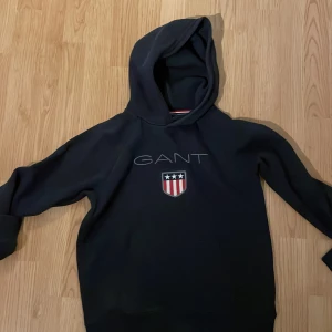 Svart hoodie från GANT Pris går att diskutera - Säljer en snygg blå hoodie från GANT. Den har en klassisk design med GANT-loggan och ett sköldemblem på framsidan. Perfekt för en casual look och passar bra till både jeans och joggers. Den är i bra skick och redo för en ny ägare!
