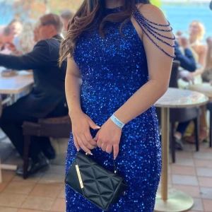 Blå glittrig klänning - Säljer en fantastisk blå glittrig klänning som verkligen fångar ögat! Den har en tight passform och är perfekt för festliga tillfällen. Klänningen är axelbandslös med snygga kedjedetaljer över axlarna. Perfekt för en kväll ute eller en speciell fest!