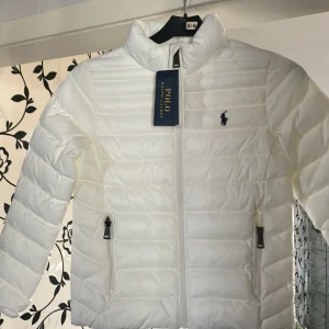 Vit pufferjacka från Polo Ralph Lauren - Säljer en snygg och stilren vit pufferjacka från Polo Ralph Lauren. Jackan har en quiltad design med dragkedja framtill och två praktiska fickor med dragkedjor. Perfekt för kyliga dagar och ger en sportig look. Den är i nyskick och redo för nya äventyr!