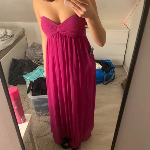 Rosa axelbandslös långklänning - Säljer en snygg rosa axelbandslös långklänning med en knutdetalj framtill. Perfekt för sommarens festligheter eller en elegant kväll. Klänningen har en flytande passform som ger en vacker siluett.