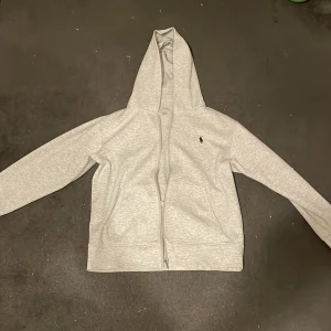 Grå hoodie från Ralph Lauren - Säljer en stilren grå hoodie från Ralph Lauren med dragkedja och det klassiska logotypbroderiet på bröstet. Perfekt för en avslappnad look. Passar bra till bland annat jeans.