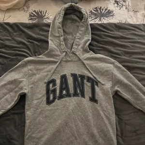 Grå hoodie från GANT - Säljer en stilren grå hoodie från GANT med stor logga på bröstet. Tröjan har en klassisk huva med dragsko och långa ärmar. Perfekt för en avslappnad look.