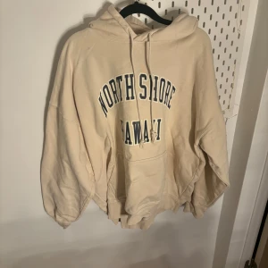 Beige hoodie med text - Säljer en beige hoodie med texten 'North Shore Hawaii' tryckt på framsidan. Den har en rymlig passform med en stor ficka framtill och en justerbar huva. Perfekt för en avslappnad stil.
