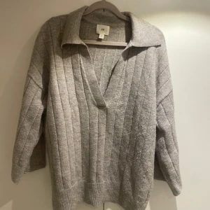 Grå ribbad tröja från H&M - Säljer en stilren grå ribbad tröja från H&M med en djup v-ringning och krage. Tröjan har trekvartsärmarär och en avslappnad passform, perfekt för kyligare dagar. Knappt använd 