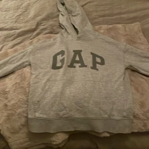 Grå hoodie från GAP Kids - Säljer en klassisk grå hoodie från GAP Kids i storlek  (12). Tröjan har en stor GAP-logga på framsidan och är tillverkad i en mjuk bomullsblandning med huva och långa ärmar. Perfekt för en avslappnad stil.