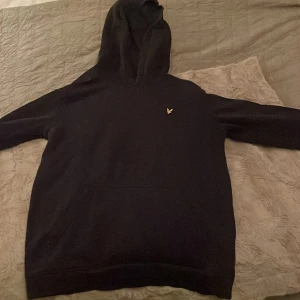 Marinblå hoodie från Lyle & Scott - Säljer en stilren svart hoodie från Lyle & Scott Junior. Den har en klassisk design med en liten gul logga på bröstet och en bekväm huva. Perfekt för en avslappnad look.