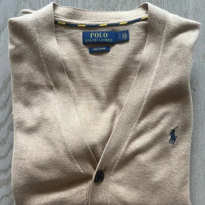 Beige kofta från Ralph Lauren - Säljer en stilren beige kofta från Ralph Lauren i mjuk pima bomull. Koftan har en v-ringning och knappar framtill samt två fickor. Den ikoniska loggan är broderad på bröstet. Perfekt för en avslappnad och elegant look.
