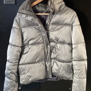 Grå pufferjacka - Säljer en stilren grå pufferjacka med dragkedja och knappar. Jackan har långa ärmar och en hög krage för extra värme. Perfekt för kyliga dagar. Liknar Henro och Massimo dutti jackan💕