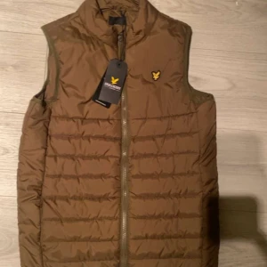 Olivgrön dunväst från Lyle & Scott - Säljer en olivgrön dunväst från Lyle & Scott med dragkedja och broderad logga på bröstet. Perfekt för kyligare dagar och ger en stilren look. Västen är lätt och bekväm att bära. helt ny är den använd en gång