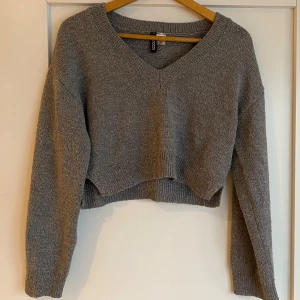 Grå stickad tröja från H&M Divided - Säljer en grå stickad tröja från H&M Divided. Tröjan har en croppad design med v-ringning och långa ärmar. Perfekt för en stilren och bekväm look. Passar bra till både jeans och kjol. Tröjans färg är som på bild 3.