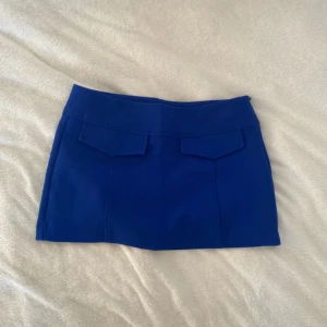 Blå kjol från Pull&Bear - Snygg blå kortkjol från Pull&Bear. Kjolen har en stilren design med två dekorativa fickor framtill. Perfekt för en trendig look.