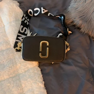 Svart och vit axelväska från Marc Jacobs - Snygg axelväska från Marc Jacobs i svart och vitt med guldiga detaljer. Väskan har en bred axelrem med logotyp och en praktisk dragkedja. Perfekt för att bära dina nödvändigheter med stil.