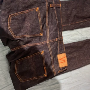 Mörkblå jeans från Nudie Jeans - Snygga mörkblå jeans från Nudie Jeans med orange sömmar. Klassisk femficksdesign och knappgylf. Perfekta för en stilren look.